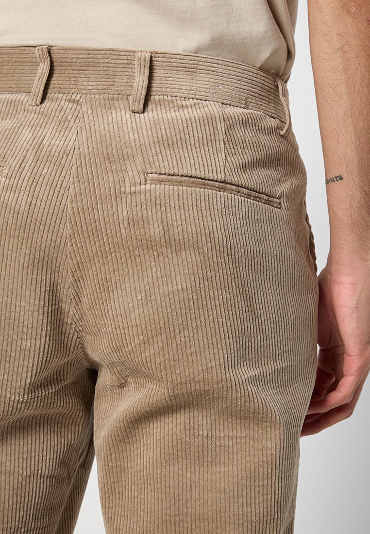 Beige Cordhosen mit Gürtelschlaufen und einer Gesäßtasche auf einer Person, die ein beiges Hemd trägt, rechte Hand seitlich sichtbar.