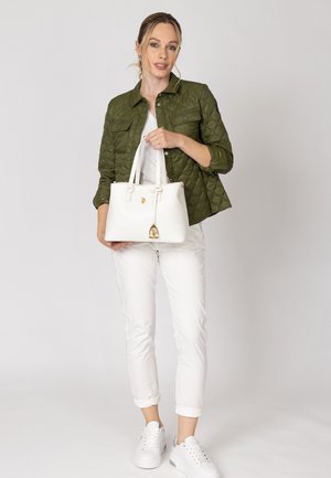 Femeie purtând o geacă matlasată verde măsliniu și pantaloni albi, ținând o geantă albă cu accente aurii, stând pe un fundal deschis.