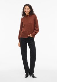 Kvinna med lockigt hår iförd en rostfärgad tröja, svarta jeans och svarta klackstövlar, stående och leende mot en vit bakgrund.