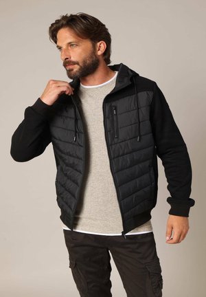 Homme barbu aux cheveux bruns portant une veste matelassée noire sur un pull beige et un pantalon cargo foncé, ajustant le col de la veste.