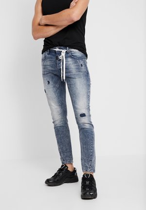 Vaqueros de denim de color azul claro con detalles desgastados, corte ajustado y tobillos remangados. Combinados con una blusa sin mangas negra y zapatillas negras.