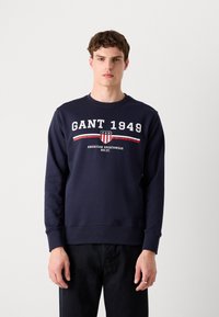Marine sweatshirt met het "GANT 1949" logo, witte en rode accenten, ronde hals en geribbelde boorden. Model draagt een donkere jeans.