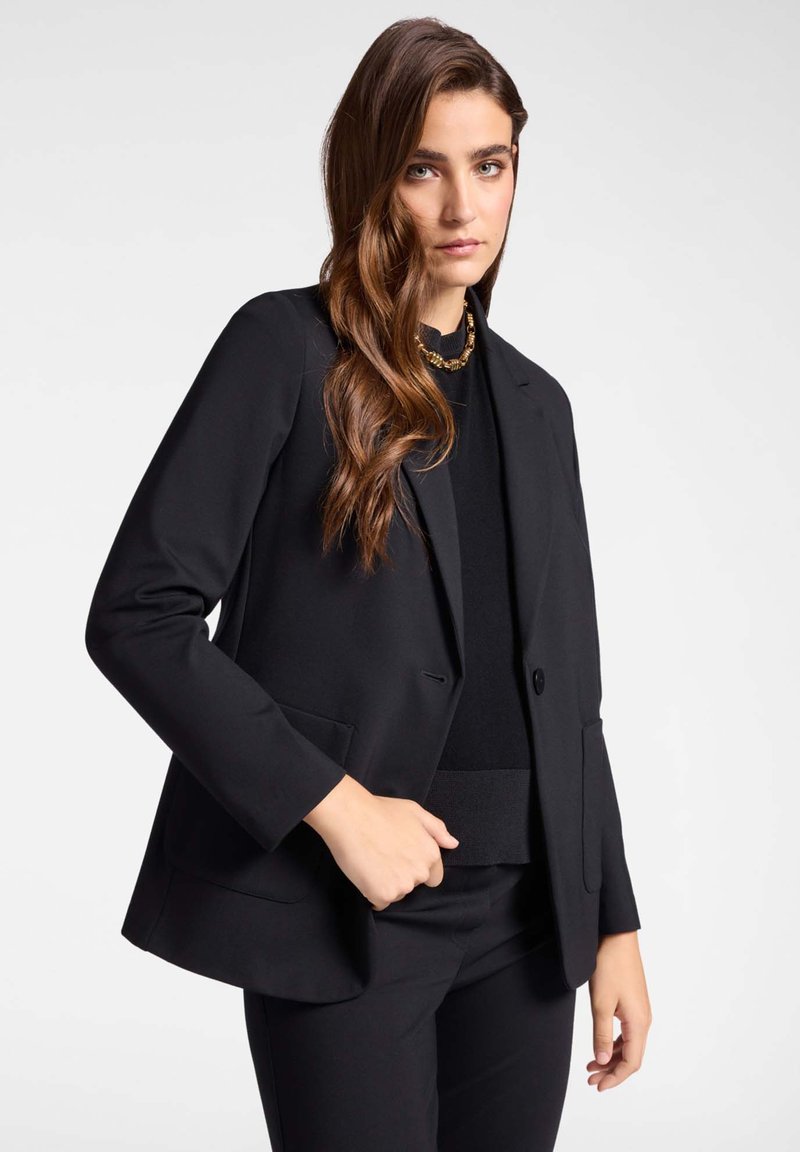Blazer nero su misura con rever, chiusura a un bottone e due tasche frontali. Indossato sopra una maglietta nera, caratterizzato da una texture di tessuto liscia.