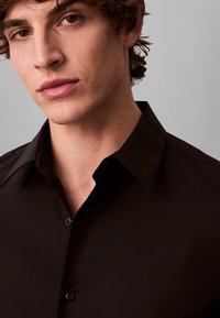 Black button-up shirt with a collared design. Features a subtle embroidered logo on the left chest. Smooth texture and tailored fit.