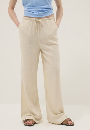 Vrouw die beige wijde broek met trekkoord in de taille en bruine sandalen draagt, staand met handen in de zakken tegen een effen achtergrond.