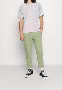T-shirt pastel légèrement motifé, avec des tourbillons en rose, bleu et blanc, associée à un pantalon de survêtement vert ajusté et des baskets noires.