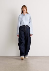 Polo a maniche lunghe a righe blu chiaro abbinato a jeans scuri a vita alta. Modello che indossa sneakers color crema, in piedi di fronte a un muro neutro.