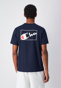 Camiseta de algodón azul marino con un logo de Champion en blanco y rojo en la parte trasera dentro de un contorno rectangular. Diseño de mangas cortas y cuello redondo.