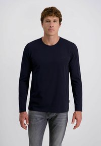 Mann mit lockigem braunem Haar, der ein enges schwarzes Langarmhemd und graue Jeans trägt, steht vor einem schlichten weißen Hintergrund.