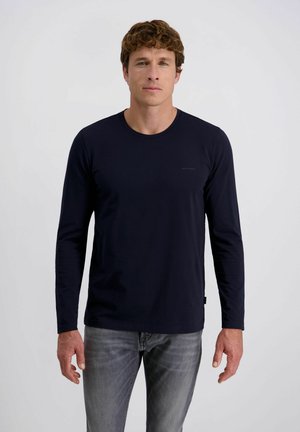Man met krullend bruin haar, gekleed in een getailleerd zwart longsleeve-shirt en grijze jeans, staand tegen een effen witte achtergrond.