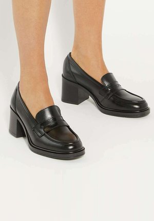 Mocassins en cuir noir usés à talons blocs, bouts arrondis et détail de bride sur une surface blanche lisse.