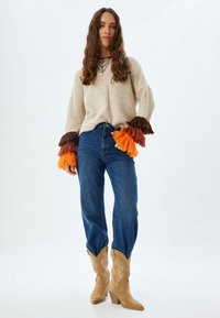 Maglione beige lavorato a maglia con polsini decorati da frange colorate in marrone, arancione e rosso. Jeans blu a vita alta e stivali da cowboy color sabbia con bordo curvo.