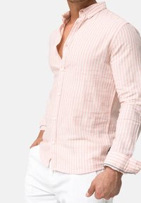 Mann in einem hellrosa und weißen vertikal gestreiften Hemd mit hochgekrempelten Ärmel und weißen Hosen, Hände in den Taschen.