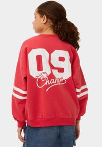 Felpa rossa con grande stampa bianca "09" e logo cursivo "Champs" sul retro, con strisce bianche su ciascun manica. Tessuto morbido, vestibilità over.
