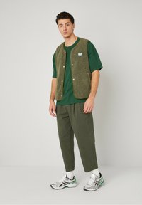 Green fleece vest with snap buttons over a dark green t-shirt, paired with loose olive green pants and silver athletic sneakers.