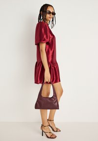 Κόκκινο σατέν φόρεμα με φουσκωτά μανίκια και ραμμένο στρίφωμα. Συνδυάζεται με μπορντό τσάντα χειρός και πέδιλα με λουράκια. Πλεξούδα και γυαλιά ηλίου.