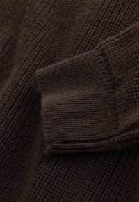 Brown knitted sweater with ribbed texture, featuring a folded cuff. The fabric appears soft and thick, showcasing varying stitch patterns.
