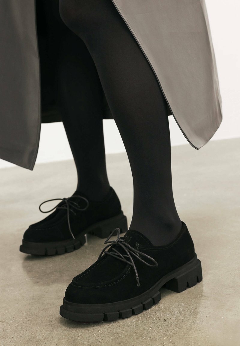 Pantofi din suede negru cu talpă groasă și cu prinderi din material. Designul prezintă o vârf rotunjit și detalii de cusătură vizibile.