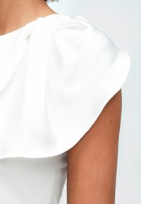 Woman wearing an off-shoulder white satin dress with a large ruffle detail over the shoulder against a plain background.