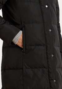 Melnā pūkāmā mēteli ar quilted dizainu, pogām ar spiedpogu un sānu kabatu, ar pelēku ribotu trikotāžas aproci, kas redzama piedurknē.