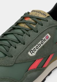 Zelena Reebok tenisica sa gornjim dijelom od antilopa i mrežaste tkanine, ružičasti detalji, istaknuta oznaka logotipa i dizajn s vezicama. Vidljiva crna gumena obloga.