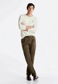 Νεαρός άνδρας που στέκεται με τα χέρια σταυρωμένα, φορώντας άσπρο μακρυμάνικο πουκάμισο, ελαιόχρωμο παντελόνι και καφέ παπούτσια σε λευκό φόντο.