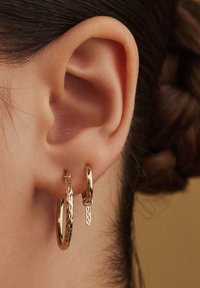 Boucles d'oreilles en or affichées sur l'oreille ; un gros anneau texturé et un petit anneau lisse avec un accent pendentif. Surface brillante, formes circulaires.
