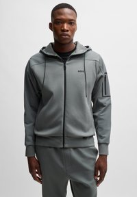 Grijze zip-up hoodie met verstelbare capuchon, zwarte accenten en logo op de borst. Bevat zijzakken en een gestructureerde stof in een sportief ontwerp.