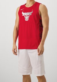 Camiseta de baloncesto roja con un gráfico de un toro en blanco, con un cuello redondo y un diseño lateral en negro. Combinada con pantalones cortos de malla blancos.