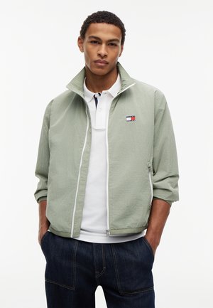 Ung mand iført lys grøn lynlåstrøje over hvid polo og mørke jeans, stående med hænderne i lommerne mod en hvid baggrund.