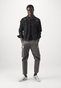 Chaqueta de denim negra con acentos desgastados, camiseta negra, pantalones de chándal grises con rayas azules y logotipos, y zapatillas blancas con rayas negras.