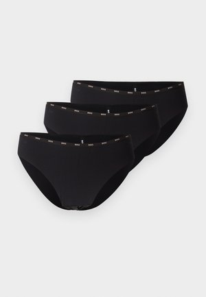 Femme debout pieds nus portant un soutien-gorge beige clair et des sous-vêtements noirs sur un fond gris clair uni.