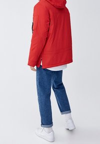 Persona che indossa una giacca a vento rossa con cappuccio, jeans blu arrotolati, calzini bianchi e sneaker bianche, in piedi contro uno sfondo chiaro e semplice.
