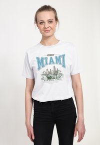 Hvid bomulds T-shirt med et grafisk design, der har teksten "NÆSTE STOP MIAMI" i teal, der viser Miami skyline og palmeblade.
