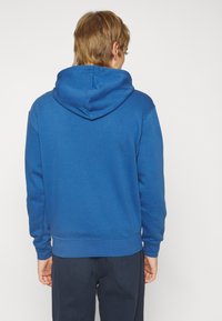 Sudadera azul con capucha, hecha de una tela suave, que presenta un bolsillo tipo canguro y puños acanalados. La vista trasera muestra un ajuste relajado y un diseño sin costuras.
