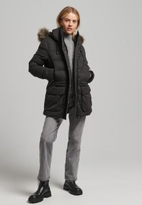 Czarna kurtka puchowa z kapturem wyłożonym futrem, z przednimi kieszeniami, z zamkiem błyskawicznym i regulowanymi sznurkami. W połączeniu z szarymi dżinsami i czarnymi botkami.