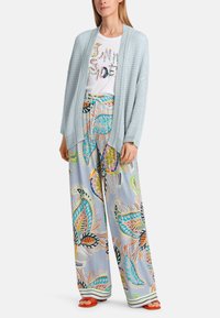 Helles blauer Strickcardigan über einem weißen Grafik-T-Shirt und bunten, gemusterten weiteren Hosen mit Fisch- und Blumendesigns. Orangefarbene Slides.