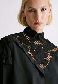 Blusa nera con alto colletto arricciato, pannello in pizzo floreale elaborato e maniche corte a palloncino, caratterizzata da una texture morbida e un design elegante.