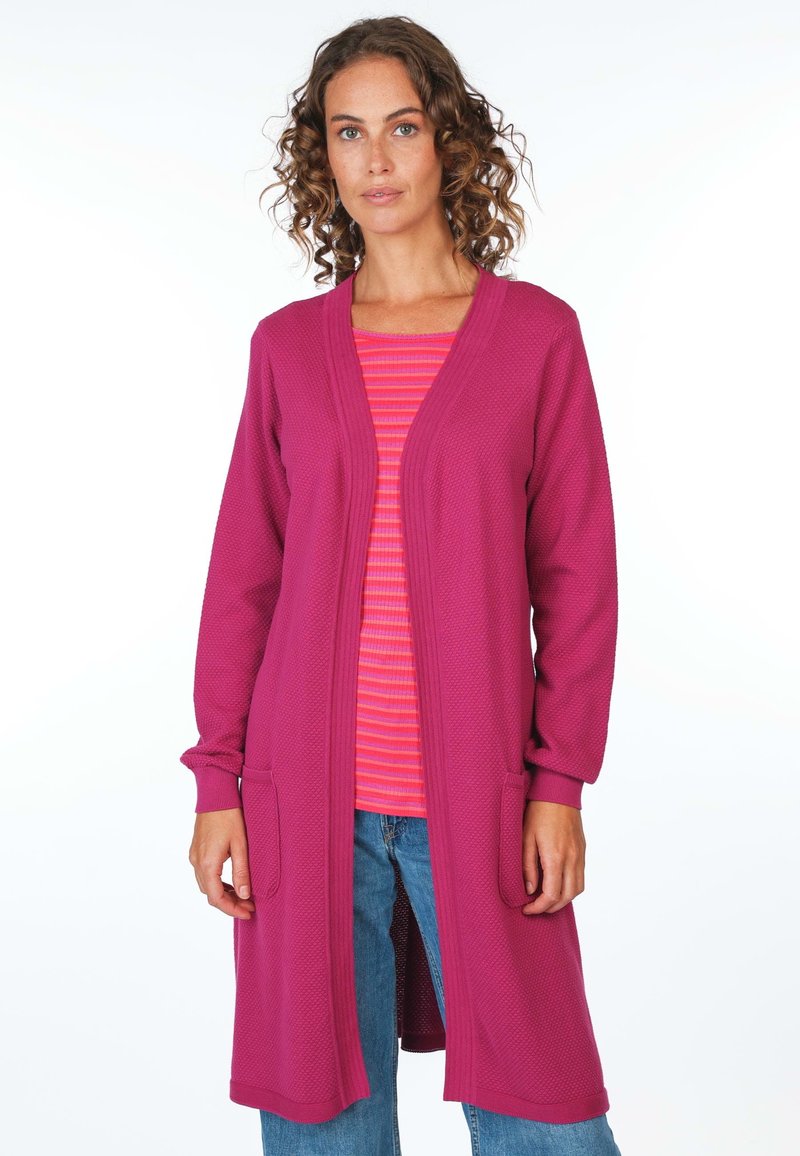 Langer, offener Cardigan aus strukturiertem fuchsiafarbenem Stoff mit Taschen. Getragen über einem gestreiften rosa Oberteil, kombiniert mit blauen Jeans.