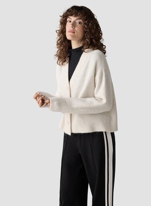 Frau mit lockigem Haar, die eine weiße, geknöpfte Strickjacke über einem schwarzen Top und schwarzen Hosen mit weißen Seitenstreifen trägt, steht vor einem grauen Hintergrund.