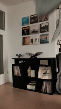 Black wooden shelf unit with vinyl records, books, a vase, and a bag. Open book on top, clock displaying 4:05, gray guitar leaning nearby.