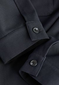 Gros plan sur des poignets de chemise en tissu noir avec des boutons noirs et des détails de couture visibles.