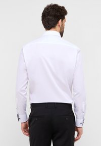 Weißes langärmliges Hemd mit strukturiertem Kragen, knopfbaren Manschetten und taillierter Passform, kombiniert mit schwarzen Hosen mit doppelten Gesäßtaschen.