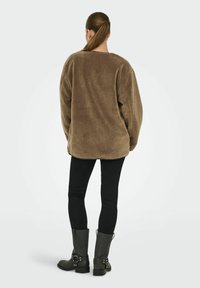 Kobieta z kucykiem, ubrana w brązową, oversize'ową kurtkę polarową, czarne legginsy i czarne botki do połowy łydki, stoi tyłem na białym tle.
