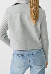 Light gray cropped jacket with a ribbed texture on sleeves and soft fabric. Worn with high-waisted light blue denim jeans.
