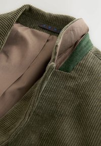 Primer plano del cuello de una chaqueta de pana verde con forro beige y un detalle de fieltro verde en el interior del cuello.