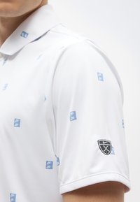 Polo blanc à manches courtes avec motif de carrés bleus au logo Nike et écusson noir Nike golf sur la manche gauche.