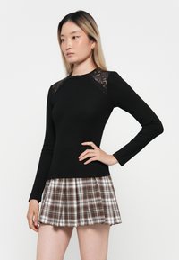 Black long-sleeve top with lace accents, paired with a pleated plaid mini skirt in white, brown, and beige tones. Smooth fabric texture throughout.