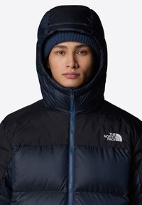 Padded czarna i granatowa kurtka z wysokim kołnierzem i kapturem, z zamkiem błyskawicznym z przodu oraz logo The North Face na piersi.