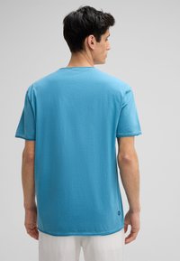 T-shirt à manches courtes, col rond, en coton bleu clair, avec des ourlets cousus et un petit logo en bas à gauche.